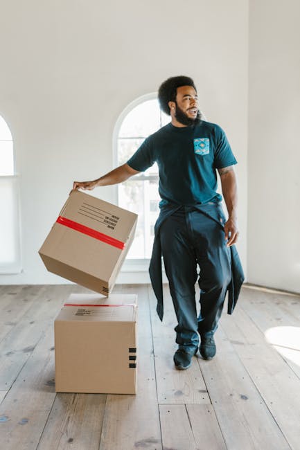 A man with dark, curly hair and a beard is inside a spacious, well-lit room with white walls and wooden flooring, holding a cardboard box with red tape and black barcode labels in one hand. He is dressed in a blue t-shirt with a blue and white logo on the chest, dark trousers, and has a jumper tied around his waist. In front of him, two additional cardboard boxes are stacked on the floor, one of which also has red packing tape and barcode labels. The large window behind him allows natural light to fill the space, highlighting the preparation and packing phase of home relocation. This scene illustrates the process of packing and loading items for a house move, fitting the context of furniture transport and moving logistics supported by Man with Van Haringey, a professional removals service provider specializing in local moves and packing assistance.