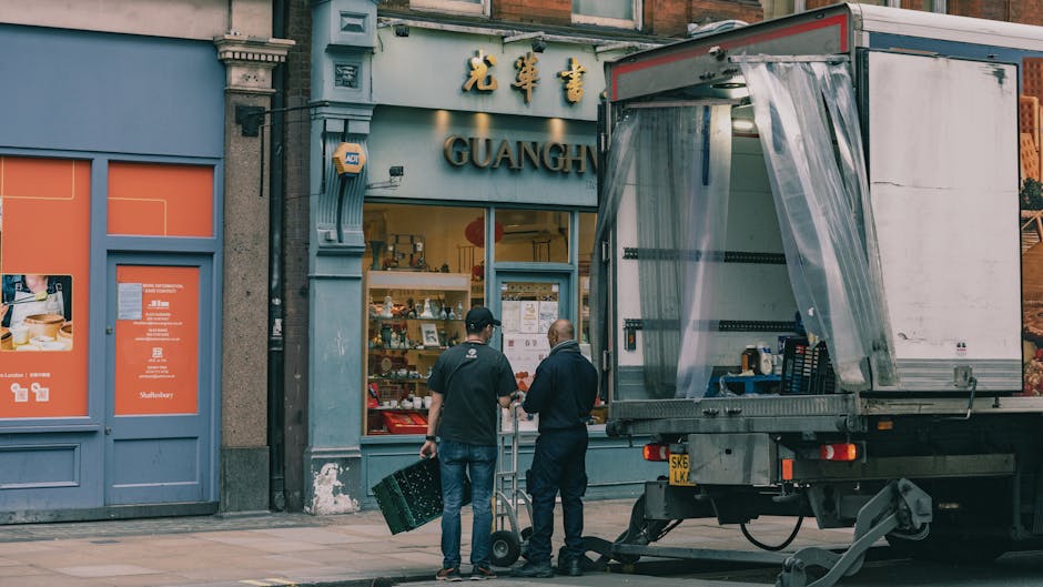 Two logistics workers in casual clothing, one with a cap, are engaged in a home relocation activity outside a shop with the sign 'GUANG' visible above the entrance, on a city street in Haringey. They are positioned near an open moving truck with a partially unfolded plastic sheet covering the cargo area, indicating an active loading process. One worker is holding a small roll of packing tape while the other manages a hand truck loaded with cardboard boxes, illustrating the packing and furniture transport phase typical of house removals. The street background features shop windows displaying various items, with a concrete façade and a blue painted wall section nearby. The scene is lit with natural daylight, capturing the practical aspects of packing, moving, and furniture logistics involved in professional removals, as carried out by [COMPANY_NAME], with emphasis on safe handling of boxes and moving equipment like hand trucks and packing blankets.