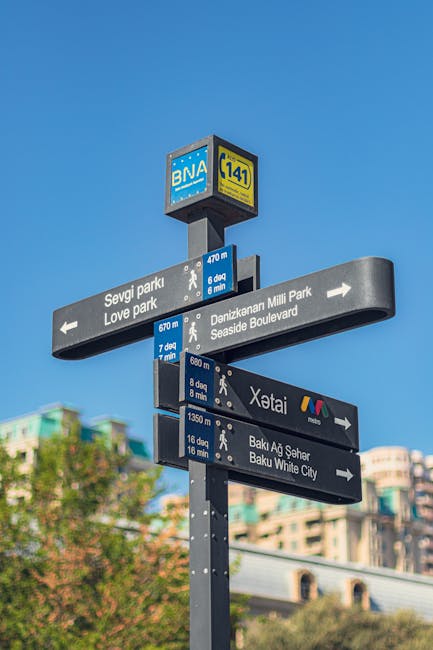 Photograph of a tall, grey directional signpost on a bright day with a clear blue sky, displaying multiple black rectangular signs with white text indicating various walking distances and directions to locations including Sevgi Park, Love Park, Denizkanar Milli Park, Seaside Boulevard, Xətai, and Baku White City. The top of the signpost features a square blue sign with the BNA logo and a yellow rectangular sign with the number 141. In the background, there are blurred multi-storey buildings and green trees, suggesting an urban area near parks and city attractions. The signpost is situated outdoors, and the signs are attached with small metal fixtures, with some signs pointing left and others pointing right, representing a navigation aid used by pedestrians. The scene emphasizes the context of location guidance within an urban environment, relevant to house removals or moving logistics in the area.