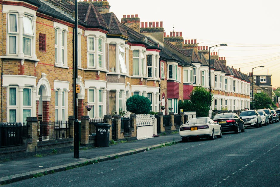 A residential street in Crouch End lined with parked cars on both sides, with the road paved in asphalt and surrounded by tall, leafy trees casting dappled sunlight onto the pavement. The street appears to be quiet, with no moving vehicles or pedestrians visible. In the background, a small traffic sign and a low fence are visible, indicating a typical suburban neighbourhood setting. The scene, captured during late afternoon or early evening, provides a calm environment suitable for home relocation activities. Man with Van Haringey often manages such streets for house removals, efficiently navigating narrow streets and managing parking restrictions during furniture transport and packing processes. The image reflects the typical environment in which professional removal services operate for house clearance and transportation within the area.