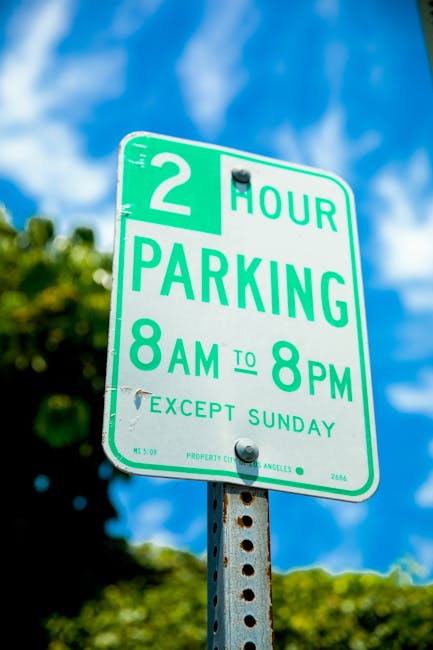A green and white parking restriction sign mounted on a metal pole with visible rust spots, indicating a two-hour parking limit from 8 am to 8 pm, Monday to Saturday, excluding Sundays. The sign is outdoors under a partly cloudy sky, with blurred green trees in the background. The sign's purpose is to inform drivers of parking rules, which is relevant for house removals and moving services like those offered by Man with Van Haringey, as they plan logistics for home relocation or furniture transport within local regulations.