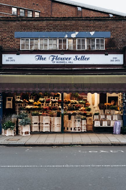 A storefront of The Flower Seller of Muswell Hill, with a brick building featuring a blue and white sign, displaying the business name and contact number. The shop has a maroon awning that partially covers the window display, which is filled with various flower arrangements, potted plants, and flower boxes. Inside the window, wooden crates and white baskets hold different types of flowers and greenery, arranged in a neat and accessible manner. The pavement in front of the shop is clear, with a curb separating it from the road, which has white road markings. The scene is captured during daytime, with natural light illuminating the floral display and the shop’s exterior, conveying a sense of a busy commercial area that may require careful planning during home relocation or furniture transport. Man with Van Haringey may assist in house removals involving similar commercial or residential properties, ensuring efficient loading and unloading during moves.