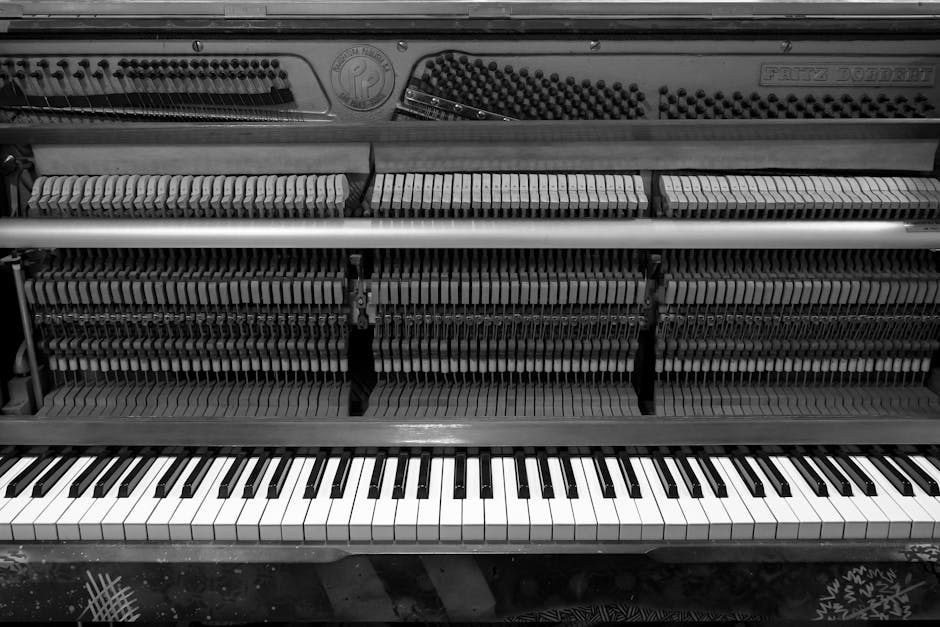 A close-up view of the interior of an upright piano with the top lid open, revealing the hammers, strings, and mechanical components in a black and white photograph. The piano's internal parts include multiple rows of hammers aligned with the strings, which are visible beneath the keys. The bottom portion shows the keyboard with black and white keys, some of which are slightly worn or discolored. The surrounding environment suggests an indoor setting with soft lighting, and the photo captures the detailed mechanisms involved in musical instrument movement and sound production. This image can support content related to packing, moving, or transporting musical instruments during home relocation or furniture transport processes, as provided by Man with Van Haringey.