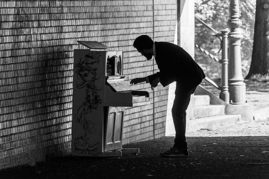 A man dressed in dark clothing, including a suit jacket and trousers, is seen outdoors leaning forward to operate an upright piano positioned against a brick wall, with his hand poised over the keys. The piano has a decorative painted design on its side and an open lid revealing the internal components. The area around him features an urban environment with a set of concrete stairs, metal railing, and a tree visible in the background, suggesting a home relocation or furniture transport scene. The scene is captured in black and white, emphasizing the focus on the manual effort involved in moving the piano, reflecting a professional move organized by Man with Van Haringey. The lighting indicates natural daylight, with shadows cast on the pavement and wall, highlighting the precise positioning and careful handling typical of professional removal services during packing and moving procedures.