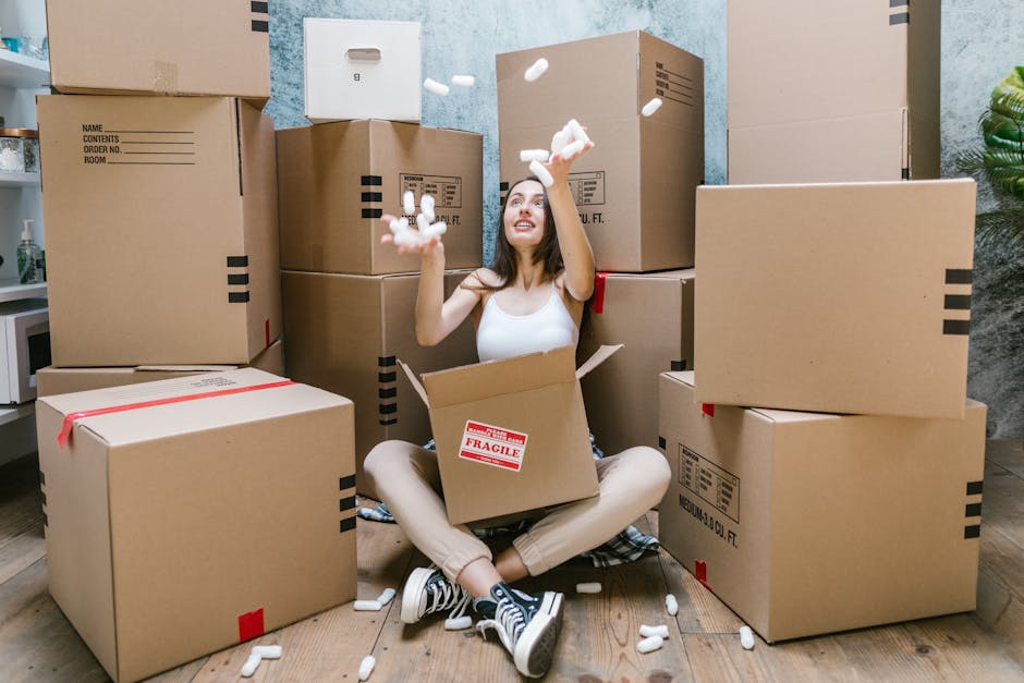 A young woman with long brown hair and casual clothing sits cross-legged on a wooden floor inside a room, surrounded by numerous large cardboard boxes, some marked with red 'fragile' labels. She is smiling and tossing white packing foam into the air, indicating a packing or moving process. The boxes vary in size and are stacked against a light blue textured wall, with some placed on top of each other and others on the floor. A red tape is visible on some boxes, and the woman is sitting next to an open box containing additional packing materials. To her left, there is a small white countertop with a bottle and other items, and to her right, a large green plant is partially visible. This scene reflects home relocation activities, including packing and furniture transport, with the environment brightly lit and organized for a household move as handled by [COMPANY_NAME], a professional removals service specializing in house and student relocations in Tottenham Hale.