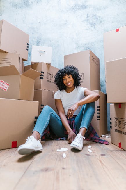 A young woman with curly black hair, wearing a white t-shirt, blue jeans, and white sneakers, is seated on a wooden floor surrounded by numerous cardboard moving boxes of varying sizes, some with shipping labels and red 'fragile' stickers. She is smiling and appears relaxed, with her arms resting on her knees. The boxes are stacked against a light blue, textured wall, indicating an indoor setting likely within a property during a home relocation or packing process. There are small white packing peanuts scattered on the floor around her. The environment suggests a moving or packing phase, with the woman possibly preparing for furniture transport or loading as part of a house or student removal service. The image is associated with Man with Van Haringey, a company providing reliable and affordable removals hosted on the webpage for Tottenham Hale student removals.
