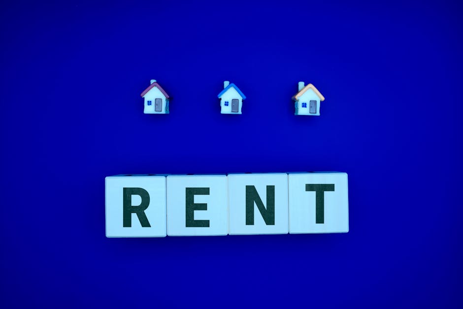 Image showing three small decorative house models with different roof colors—red, blue, and beige—mounted on a vivid blue background. Below these models is a row of four white letter blocks spelling out 'RENT' in black capital letters. The setup is arranged on a flat surface, with the houses positioned evenly above the letter blocks, which are aligned horizontally. The background is uniformly lit, emphasizing the clear contrast between the objects and the backdrop, creating a clean and organized visual related to housing, property, or rental themes relevant to house removals and relocation services offered by Man with Van Haringey.